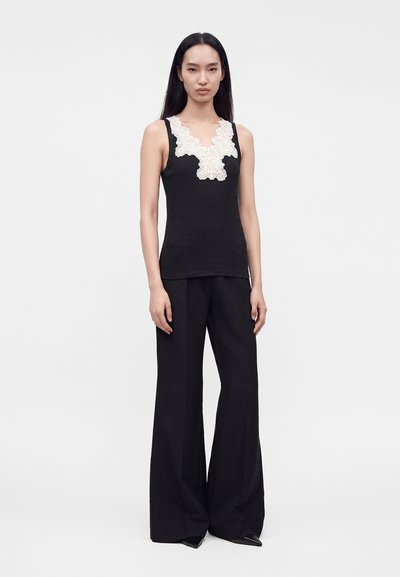 Femme aux longs cheveux noirs portant un haut noir sans manches avec des détails en dentelle blanche et un pantalon large noir sur un fond clair uni.