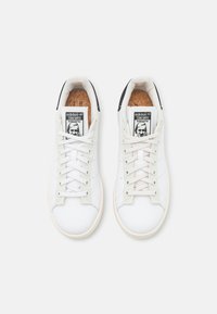 Bílé tenisky s hladkým koženým svrškem, kulatou špičkou a kontrastním černým patním panelem. Vybavené šněrováním a korkovou stélkou s detaily značky.