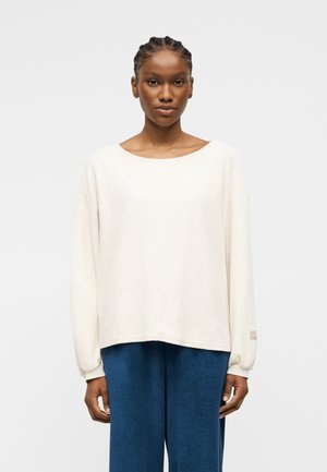 Femme aux cheveux tressés portant un haut ample à manches longues blanc et un pantalon bleu à jambes larges, debout devant un fond clair uni.