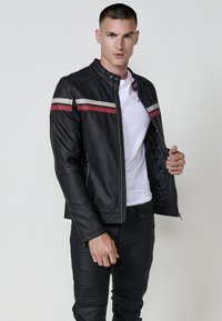 Uomo che indossa una giacca di pelle nera con strisce rosse e bianche, una maglietta bianca e pantaloni neri, che tiene aperta la giacca per mostrare l'interno.