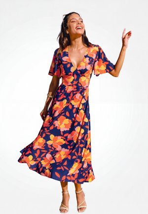 Femme souriante posant, portant une robe midi bleu marine avec un grand motif floral rose, orange et jaune, et des sandales à talons beiges.