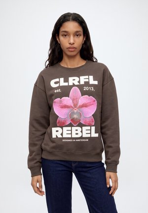 Une femme porte un sweat-shirt marron orné d’un grand motif d’orchidée rose et d’une inscription blanche en caractères gras, associé à un jean foncé.