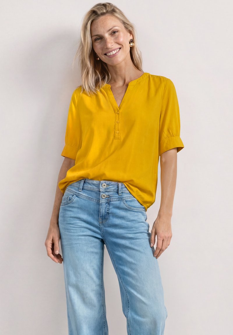 Femme souriante portant un chemisier à manches courtes jaune moutarde et un jean taille haute bleu clair sur un fond uni clair.