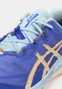 Közeli felvétel egy kék Asics FujiSpeed 4 futócipőről, világoskék fűzőkkel és bézs logóval az oldalán.