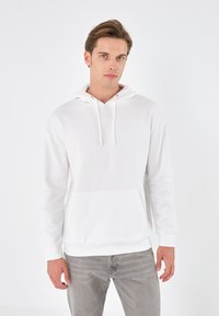 White cotton hoodie with a kangaroo pocket, drawstring hood, and ribbed cuffs. Model wears it with light grey jeans against a plain background.