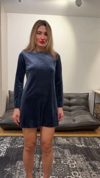 Kvinne med langt blondt hår iført en marineblå fløyelskjole med lange ermer, stående i et moderne rom med en svart skinnsofa og et mønstret teppe.