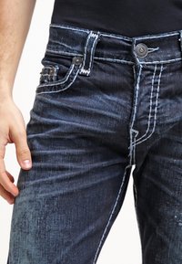 Mörkblå denimjeans med kontrasterande vit sömnad, fem fickor och metallknappar. Har en slim fit och strukturerad textil.