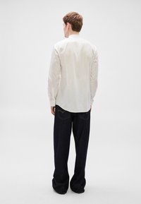 Λευκό πουκάμισο με όρθιο γιακά, μανσέτες με κουμπιά και πτυχώσεις στην πλάτη, συνδυασμένο με σκούρο μπλε τζιν παντελόνι με φαρδύ πόδι, κίτρινη ραφή και διπλές πίσω τσέπες.