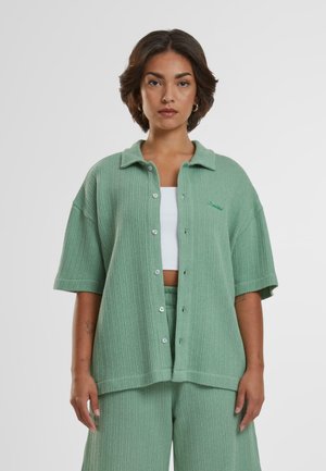 Green ribbed button-up shirt with short sleeves, featuring a collar and embroidered detail. Worn over a white top, paired with matching pants.