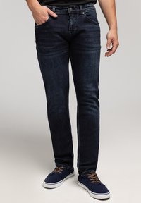 Mężczyzna w ciemnoniebieskich dopasowanych dżinsach i granatowych trampkach z brązowymi sznurówkami, z jedną ręką w kieszeni, stojący na jasnym tle.