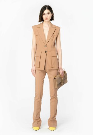 Blazer beige su misura senza maniche con due tasche frontali, abbinato a pantaloni beige aderenti e scarpe gialle a punta. Tiene in mano una borsa color sabbia.