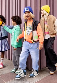 Vier kinderen in kleurrijke casual kleding en mutsen staan en interageren voor een gestructureerde paarse muur op een stedelijk trottoir.