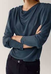Blusa drappeggiata verde acqua con maniche lunghe, dotata di pieghe sulle spalle e un collo rilassato, abbinata a pantaloni scuri e un bottone decorativo.