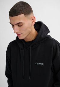 Jonge man met kort donker haar die een zwarte Hummel Sportswear hoodie draagt, kijkt omlaag tegen een effen lichtgrijze achtergrond.