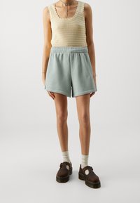 Top senza maniche in maglia beige con bordi scallop, abbinato a pantaloni corti in felpa verde chiaro a vita alta e scarpe platform marroni.
