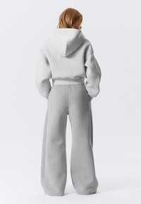 Kobieta o czerwonych włosach, ubrana w jasnoszary krótki hoodie i szerokie spodnie dresowe, stoi tyłem, ręce ma w kieszeniach.