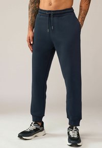 Pantalones de chándal azul marino con ajuste relajado, cinturilla elástica con cordón, bolsillos laterales y puños acanalados, combinados con zapatillas negras y grises.