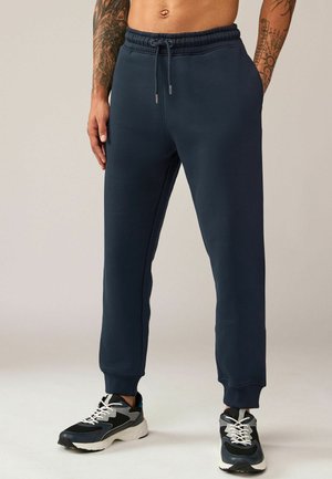 Pantaloni de trening bleumarin cu o croială lejeră, talie elastică cu șnur, buzunare laterale și mansete ribbate, asortați cu adidași negri și gri.