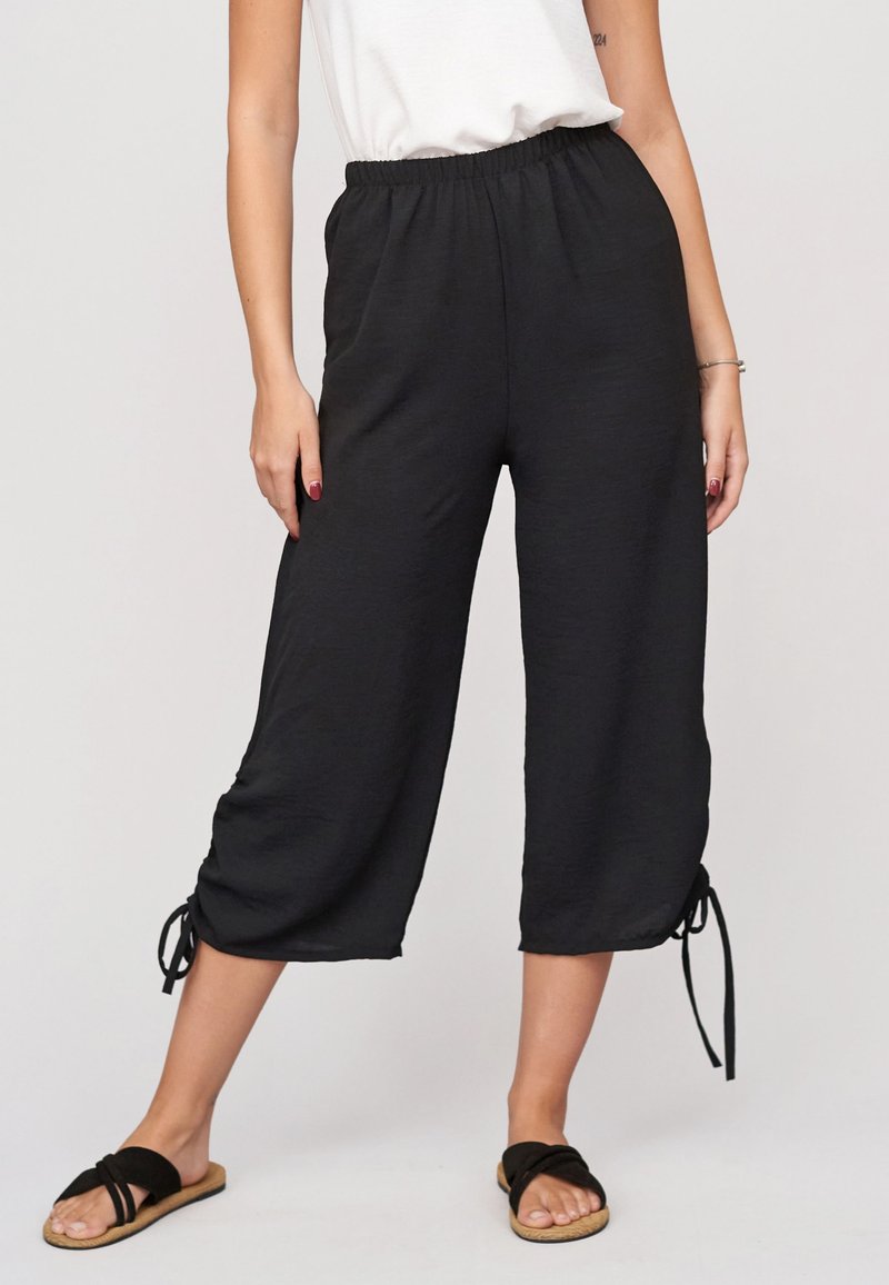 Femme portant un pantalon court noir ample avec des liens réglables aux ourlets, un haut blanc sans manches et des sandales plates noires.
