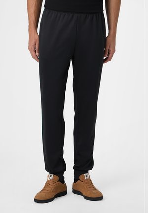 Mann trägt schwarze Jogginghose mit elastischem Bund und braune Wildledersneaker mit schwarzen Sohlen auf weißem Hintergrund.