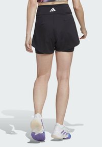 Sorte sportshorts med højt talje, med et Adidas-logo på bagsiden, parret med hvide og lilla sportssko.
