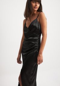 Juodas atlasinis suknelė su plonomis petnešėlėmis, giliu V formos iškirpimu ir surišimo dizainu. Yra šoninės plyšio ir susiraukšlėjimo tekstūra ant klubų, kad būtų pasiektas prigludęs efektas.