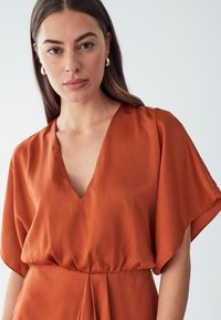 Rust-colored, silk-like blouse with a deep V-neck and flutter sleeves. Features a cinched waist for shape and a smooth texture throughout.