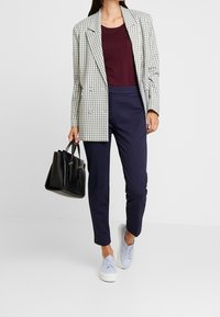 Femme portant un blazer à carreaux, un haut bordeaux, un pantalon navy, des baskets bleu clair, et tenant un sac à main noir, se tenant sur un fond blanc.