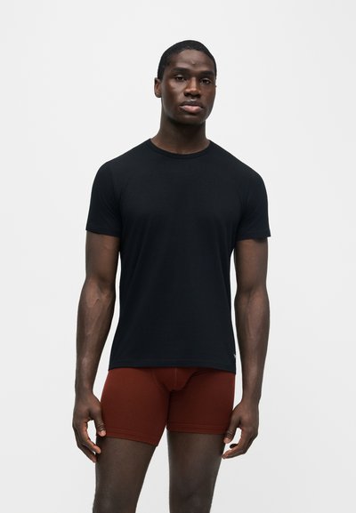 Jeune homme debout portant une chemise noire à manches courtes et un boxer couleur rouille sur un fond clair uni.