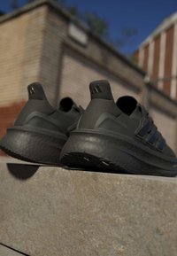 Двойка черни маратонки Adidas, почиващи на каменен парапет с тухлена сграда и синьо небе на заден план.