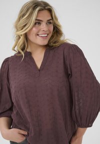 Burgundy blouse with a soft, textured finish. Features a V-neckline, short puff sleeves, and a delicate pattern of embroidered designs.