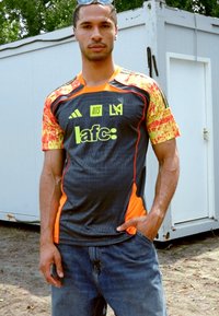 Sort og orange sportsjersey med mønstrede korte ærmer, der har logoerne af Adidas og LAFC, parret med blå jeans.