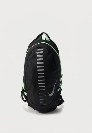 Schwarzer und grüner Nike-Rucksack mit einem vertikalen reflektierenden Streifen und Swoosh-Logo vorne, steht aufrecht vor einem einfarbigen Hintergrund.