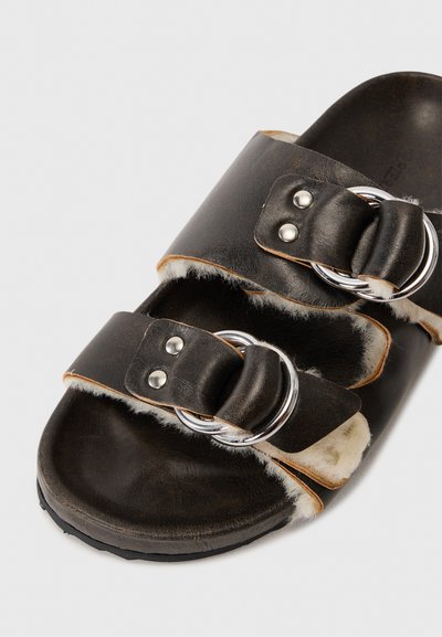 Black leather sandal with double straps, silver circular buckles, and soft white fur lining. Flat sole with textured black bottom.