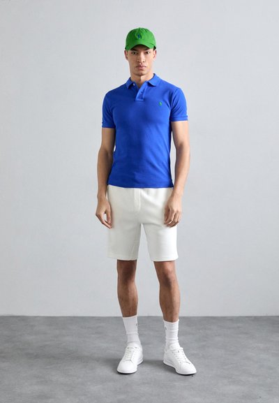 Modra polo majica, bele kratke hlače, zelena kapa. Eleganten dizajn, bombažni material, prilegan kroj. Bele superge in nogavice do gležnjev so vidne.