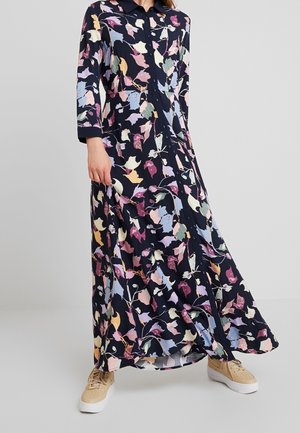Frau trägt ein langes marineblaues Blumenkleid mit Knöpfen, dreiviertel Ärmeln und beigen Sneakers, steht vor einem schlichten grauen Hintergrund.