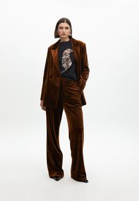 Completo in velluto marrone con giacca doppiopetto e pantaloni a gamba larga. T-shirt nera con un design di animali in paillettes e scarpe nere appuntite.