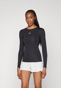 Haut de sport noir à manches longues avec un col rond et un logo blanc sur la poitrine, associé à un short clair avec un logo.