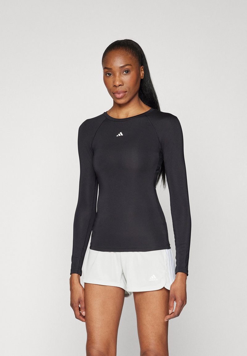 Haut de sport noir à manches longues avec un col rond et un logo blanc sur la poitrine, associé à un short clair avec un logo.