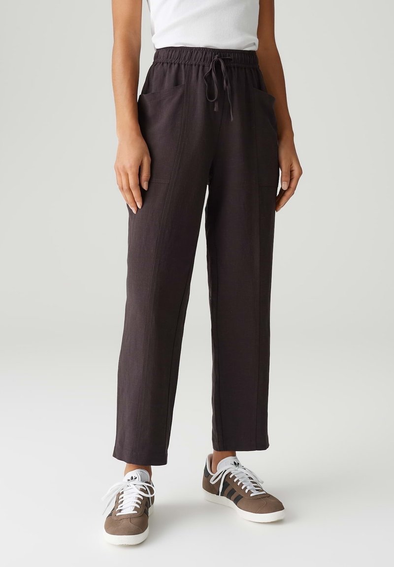 Personne portant un pantalon ample de couleur foncée avec poches frontales, taille à cordon, baskets blanches et un haut blanc sans manches, debout sur un fond uni.