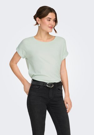 Frau trägt ein hellgrünes kurzärmeliges Hemd, schwarzen Gürtel und schwarze Jeans, steht mit einer Hand in der Hüfte und blickt zur Seite.