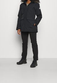 Frau, die eine schwarze Adidas-Hoodie-Jacke, schwarze Hose und schwarze Wanderschuhe trägt, steht vor einem schlichten Hintergrund.