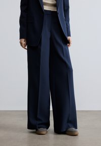 Navy wide-leg trousers made of smooth fabric, featuring a relaxed fit and subtle pleats, paired with gray sneakers for a casual look.