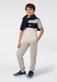 Chłopiec stoi z rękami w kieszeniach dresów, ubrany w kolorowy t-shirt Champion, beżowe dresy cargo, granatowy sweter na ramionach i niebieskie trampki.