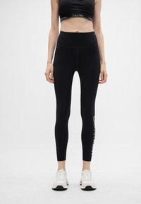 Fekete leggings magas derékkal, a bal lábszáron nyomtatott "Calvin Klein" logóval. Sima textúra, testhez simuló fazon. Fehér sportcipők.