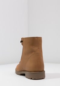 Bottine en cuir marron avec une texture lisse, des coutures minimales et une semelle en caoutchouc. Dotée d'une fermeture éclair sur le côté et d'une petite sangle décorative.