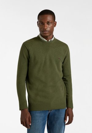 Groen gebreid sweater met een ribbelronde halslijn en manchetten, met een klein logo accent op de borst. Gepaard met blauwe jeans.