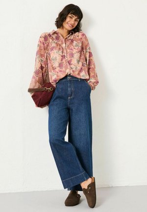 Fliederfarbende Bluse mit langen Ärmeln, kombiniert mit weiten blauen Jeans und braunen Wildlederschuhen. Eine burgunderrote Handtasche vervollständigt das Outfit.