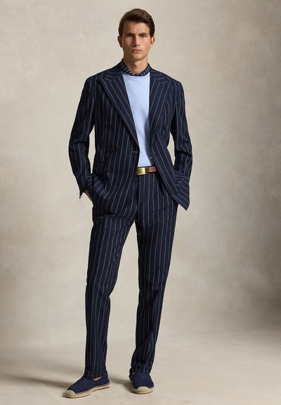 Homme debout les mains dans les poches, portant un costume bleu marine à rayures, une chemise bleu clair, une écharpe sombre, une ceinture marron et des espadrilles bleu marine.