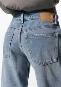 Jeans iz denima v svetlo modri barvi, z visokim pasom, standardnimi žepi in svetlo rjavim usnjenim žigom na hrbtnem pasu. Opažna je subtilna bledica.